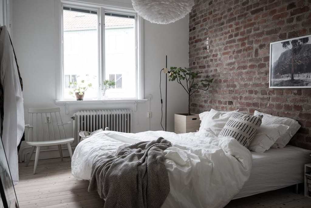 A bedroom with exposed brick and white bedding, white dry wall by the window