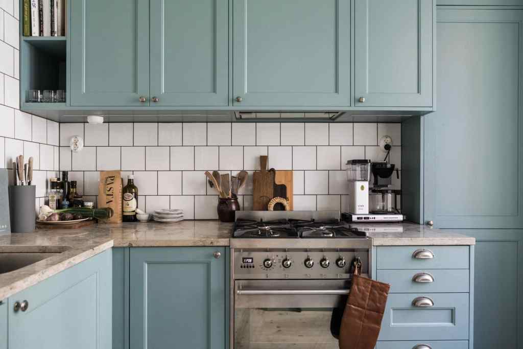 Pale blue kitchen cabinets paired with beige limestone countertops, stainless steel appliances and a white subway tile backsplash