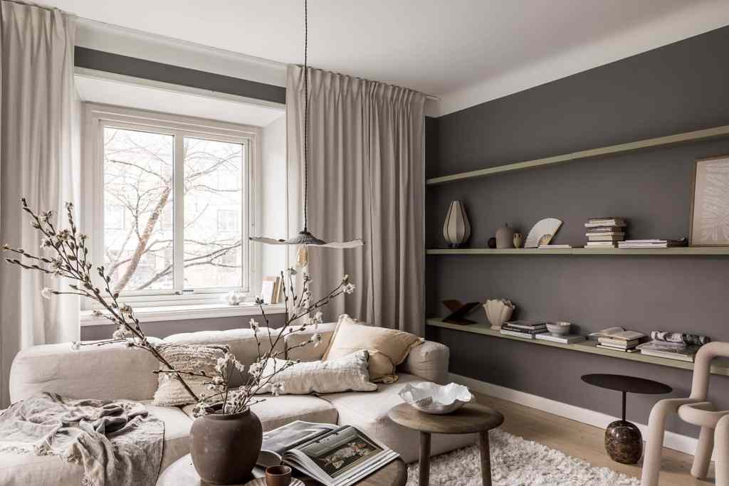 A living room with dark grey walls, a beige fabric sofa and floating shelves on the wall