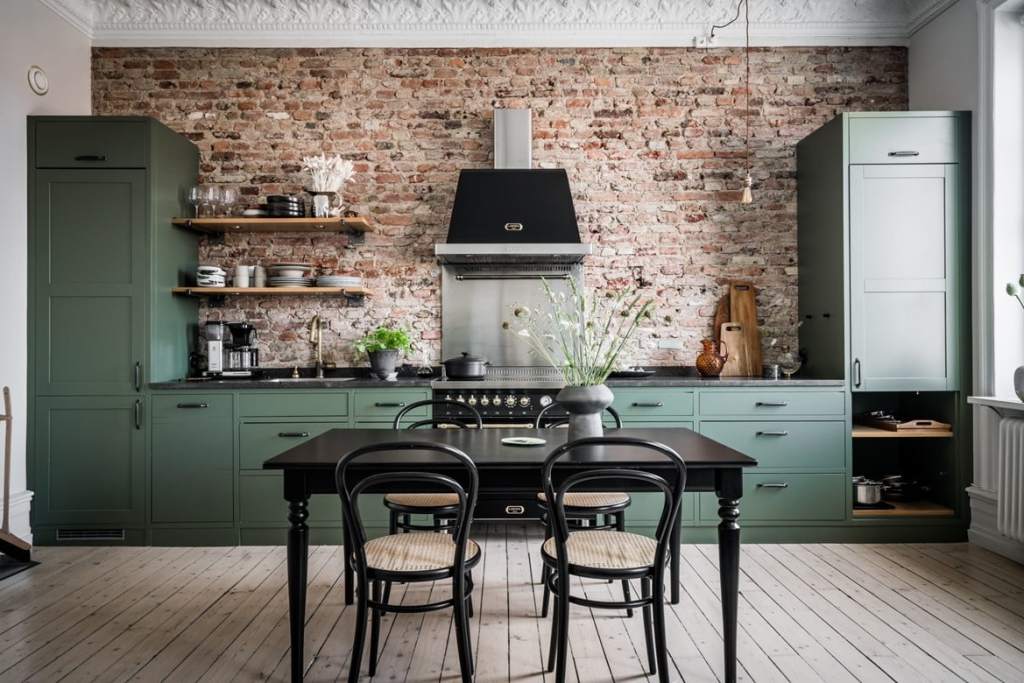 A beautiful exposed brick wall as a backsplash of a green kitchen with black countertops