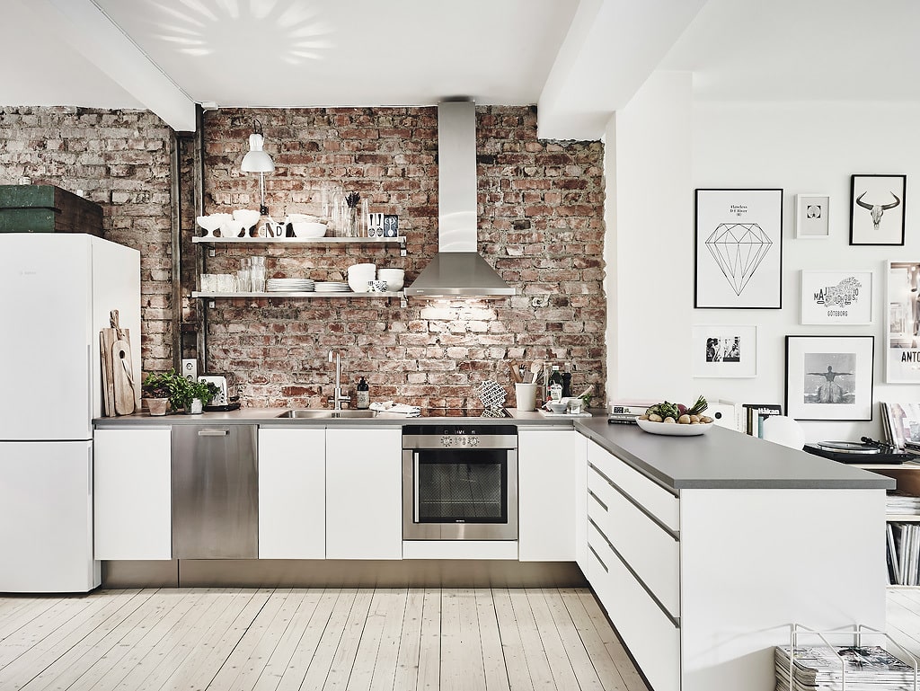 A white kitchen with grey countertops and an exposed brick wall, stainless steel appliances