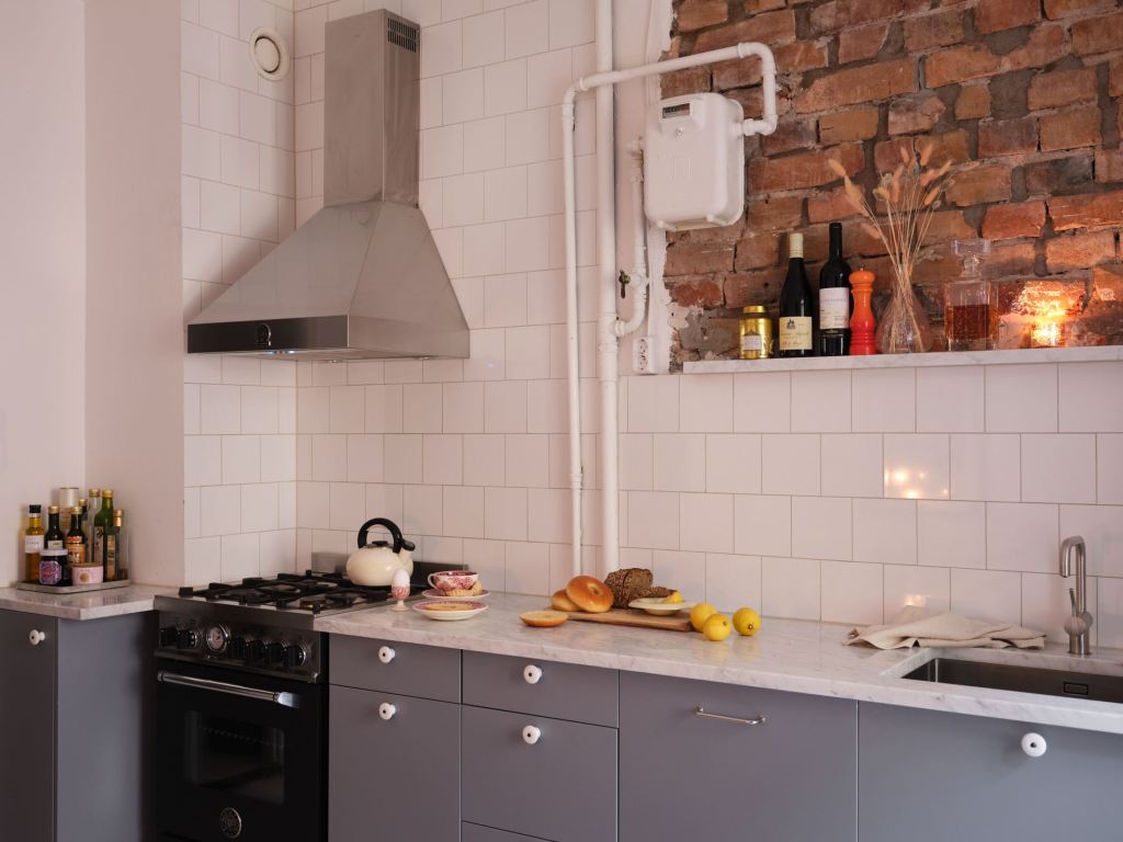 A grey kitchen with white marble countertops, a tile backsplash and exposed brick above the backsplash